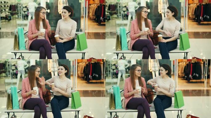 漂亮的年轻女子在商场里聊天，拿着外卖饮料坐在长椅上，纸袋站在旁边。青年生活方式和商店概念。