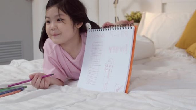 年轻的亚洲女孩在家画画。亚洲日本女儿童孩子放松休息乐趣快乐在素描本上画卡通睡觉前躺在床上，晚上在卧室