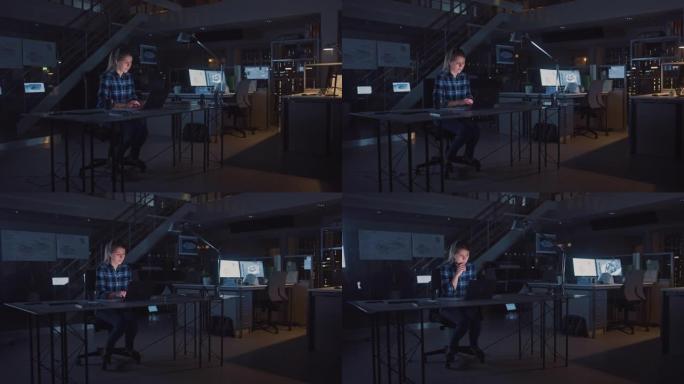 坐在办公桌前的漂亮女工程师在笔记本电脑上工作。躺在桌子上的蓝图。在黑暗的工业设计工程设施中。暖灯