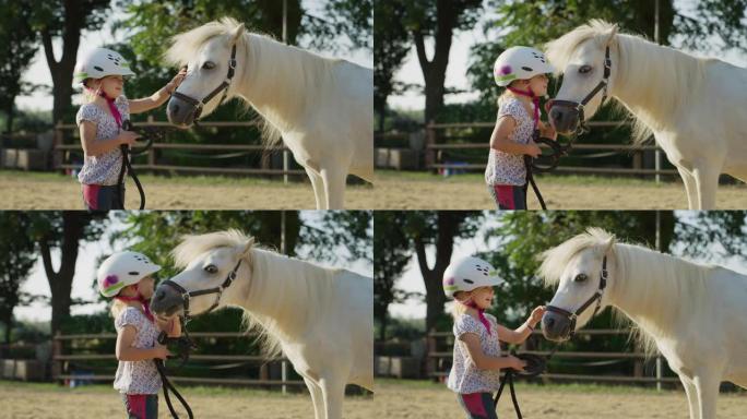 一个戴着骑师头盔的可爱的小女孩的真实镜头正在阳光下的马stable上爱抚着一匹白色的小马