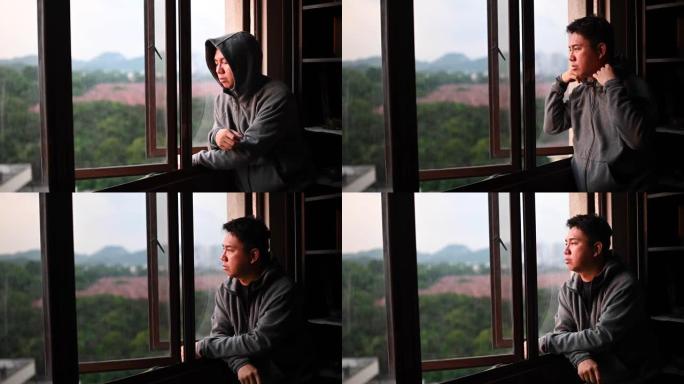 日落时分，一名身穿连帽衫的亚洲华人中年男子从家中望向窗外，表情严肃