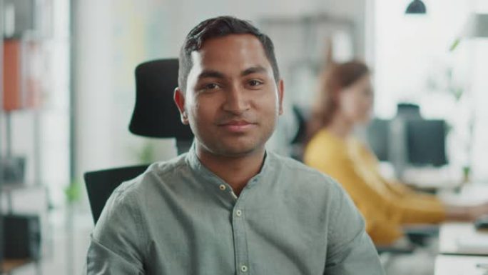英俊的职业印度男子在办公桌前工作，使用个人电脑，对着镜头微笑的肖像。在明亮多样的办公室工作的成功人士