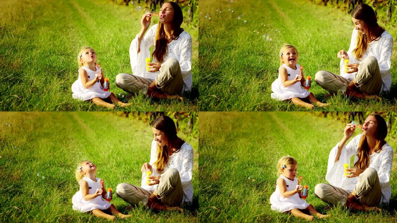 年轻母亲和婴儿2岁女孩的超级慢动作在4k的晴天在葡萄园的草地上吹气/肥皂泡