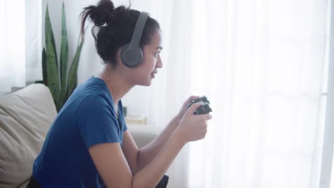 年轻的亚洲女孩玩电子游戏，在家沙发上拿着游戏控制器。
