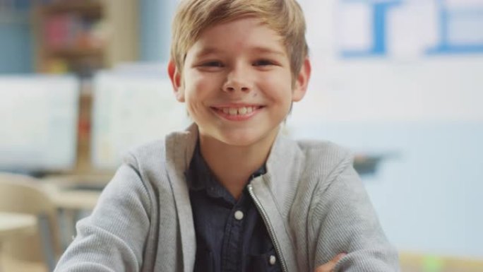 一个可爱的小男孩坐在他的课桌前，快乐地微笑着。聪明的小男孩，带着迷人的微笑坐在教室里。升降相机拍摄
