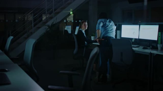 自信的商人使用台式计算机，她的男经理解释了特定的任务，帐户处理和策略。在公司办公室工作到深夜的专业人