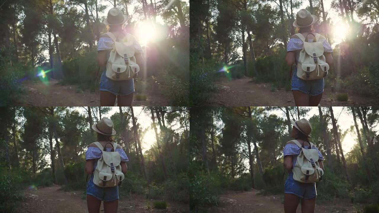 在阳光明媚的日子里，一个美丽的年轻游客 (女孩) 的肖像，背着一个背包，戴着草帽，穿过森林，树木的背