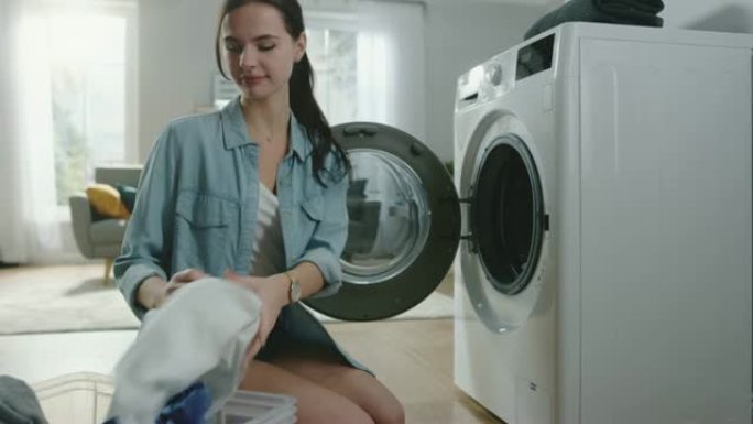 美丽微笑的黑发年轻女子穿着家常牛仔裤坐在洗衣机前。她给洗衣机装上脏衣服。明亮宽敞的客厅，室内现代。