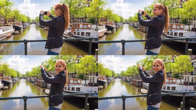 阿姆斯特丹快乐的游客，站在满是船屋的运河上的桥上，用智能手机拍照
