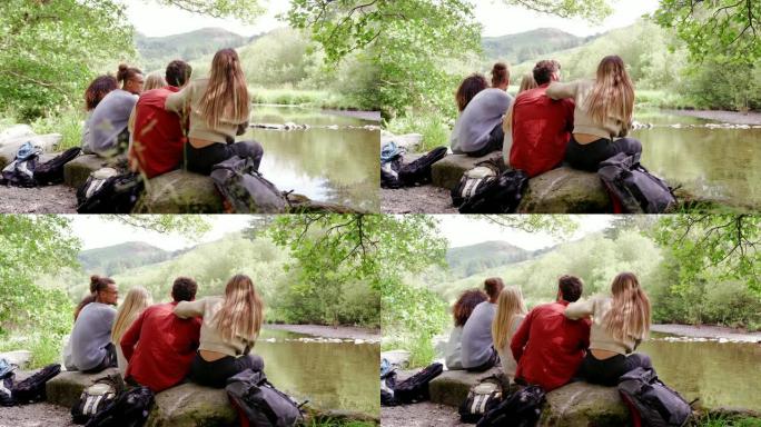 五个年轻的成年朋友在远足时坐在河边的岩石上休息时欣赏风景，向后看