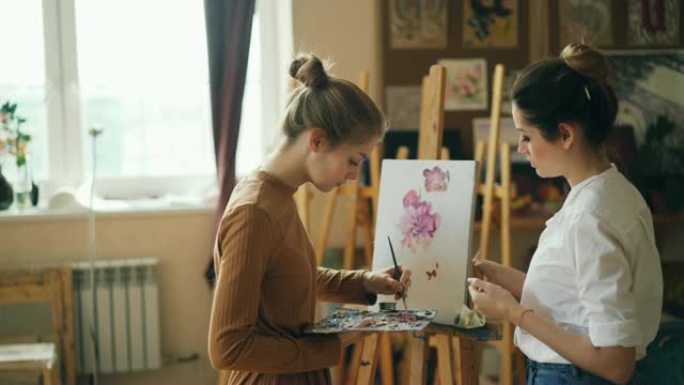 艺术学校的女学生正在用油画绘画花朵，而她的老师长得漂亮的年轻女子则在给她的建议中指向图片。