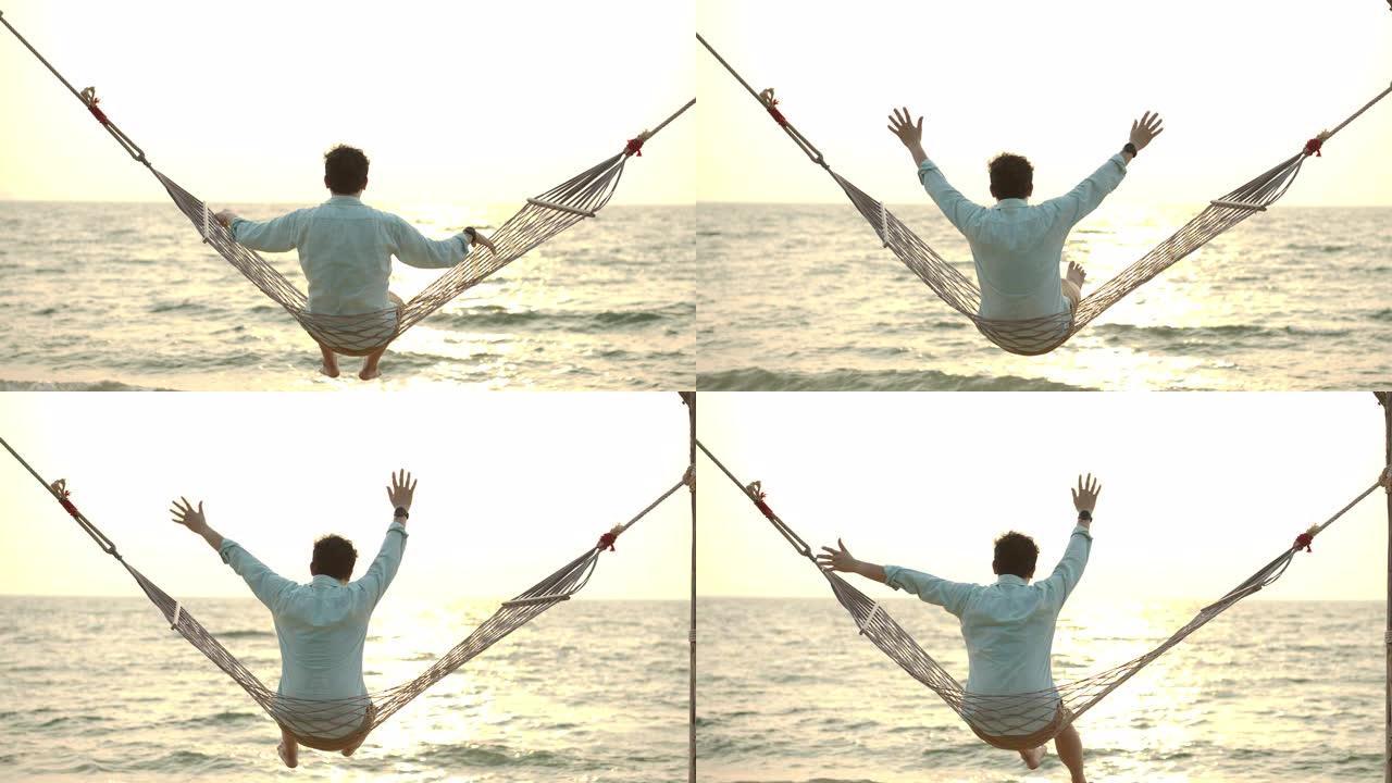 放松的男性游客享受他的暑假。年轻的亚洲男子在海滩的吊床上放松，看着地平线，欣赏日落美景。