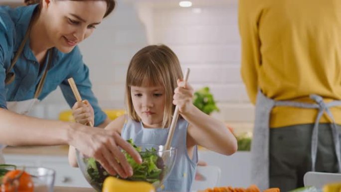 在厨房: 母亲和小女儿一起烹饪健康晚餐。妈妈教小女孩健康的习惯，以及如何在沙拉碗中混合蔬菜。可爱的孩