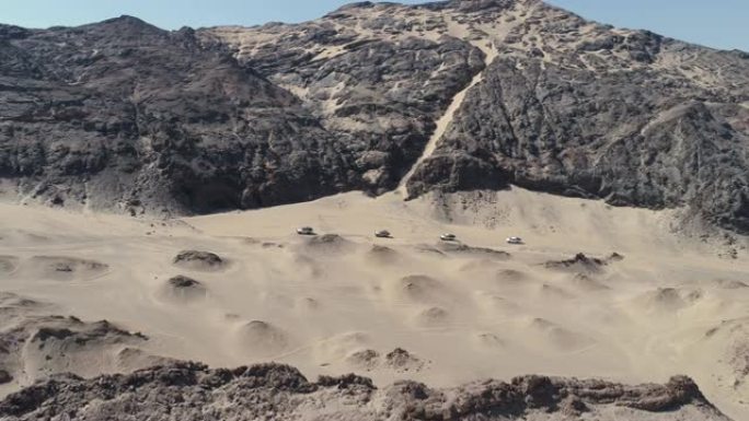 纳米比亚纳米布沙漠骨架海岸上4x4车辆车队在沙丘和山脉之间行驶的4k空中平移视图