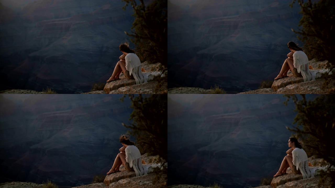 慢动作平静的年轻女子，头发飞舞，坐在雄伟的大峡谷史诗般的日落山全景上。