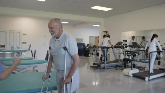 截肢者的高级男子在拐杖的帮助下与男性治疗师进行物理治疗，而其他患者则在后台锻炼