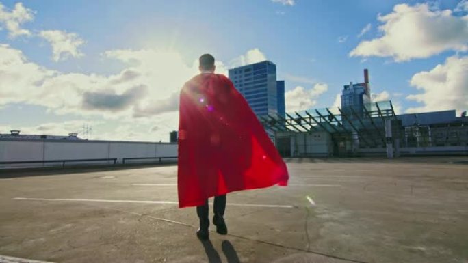 商人超级英雄，红色斗篷在风中吹动，走在摩天大楼的屋顶上，准备进行商业交易并节省一天。后视图拍摄。