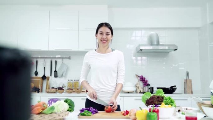 年轻的亚洲女性影响者vlogger穿着白色西装，在厨房对着镜头微笑。相机放大