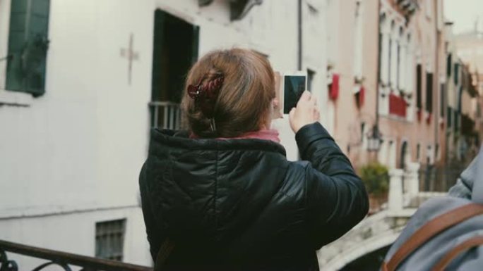 背面查看快乐的高级女旅行者拍摄意大利威尼斯美丽的秋季水渠街的智能手机照片。