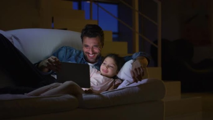 晚上在沙发上慢动作时，快乐的父亲和女儿在沙发上使用平板电脑的肖像。