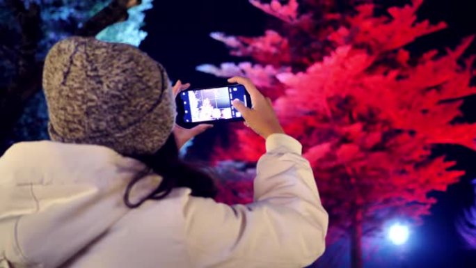 圣诞节期间，旅行者妇女用手机在圣诞树上装饰有灯光的手机拍照