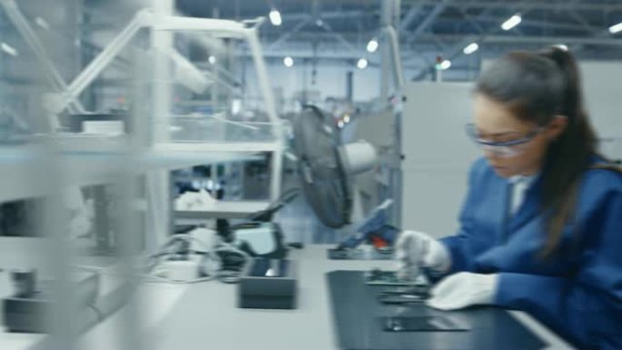 穿着蓝色和白色工作服的年轻女性和男性正在用钳子为智能手机组装印刷电路板。高科技工厂设施中的电子工厂工