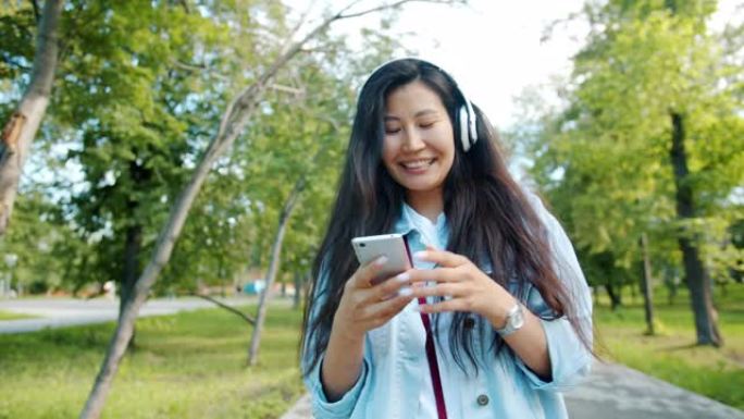 亚洲女士戴着耳机享受音乐，带着智能手机在公园散步