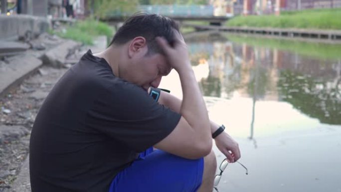 年轻沮丧的亚洲男人坐在池塘附近沮丧，悲伤，艰辛，精神卫生的观念。