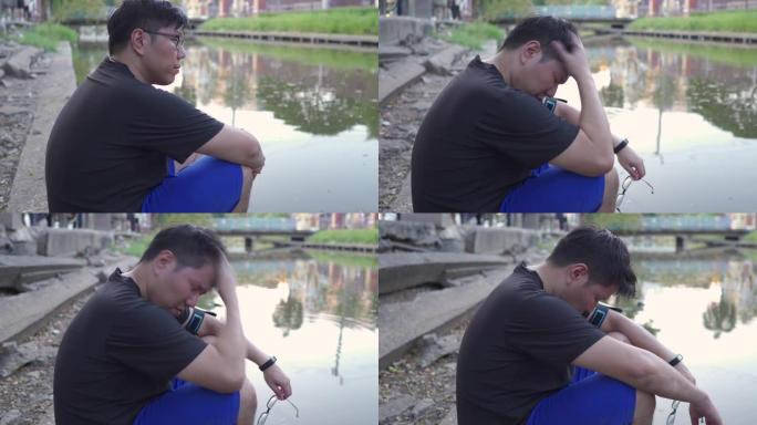 年轻沮丧的亚洲男人坐在池塘附近沮丧，悲伤，艰辛，精神卫生的观念。