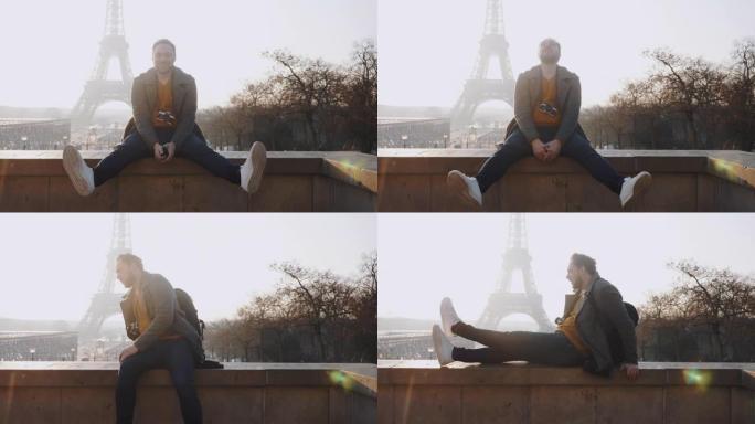 快乐的男性欧洲自由摄影师男人坐在栅栏上，微笑着看着巴黎慢动作的埃菲尔铁塔。