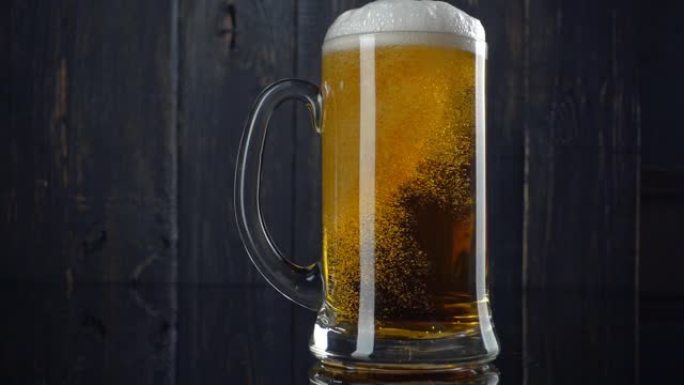 啤酒倒入啤酒杯的慢动作镜头。白色啤酒泡沫超出了杯子的边缘。陈旧的木制背景