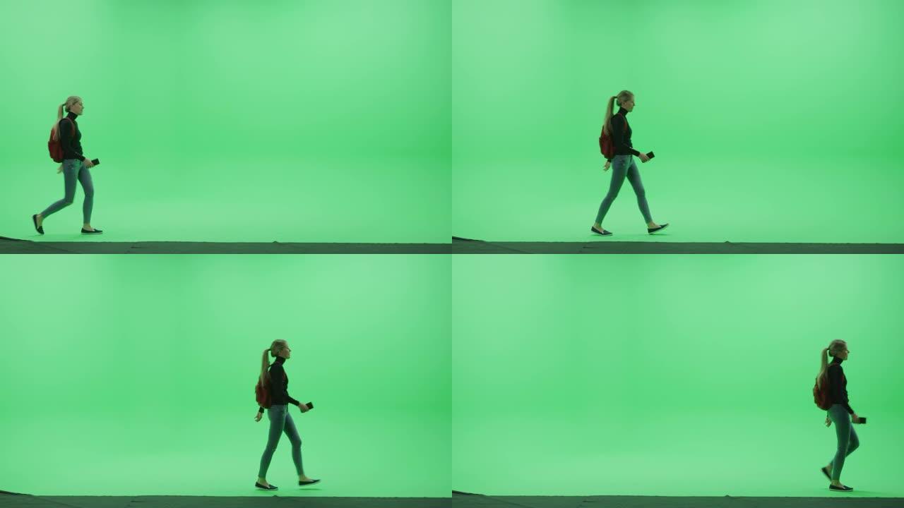 绿屏色键工作室: 美丽的年轻女子穿着时尚的高领毛衣和背包，拿着手机智能手机，从左到右愉快地走过房间。