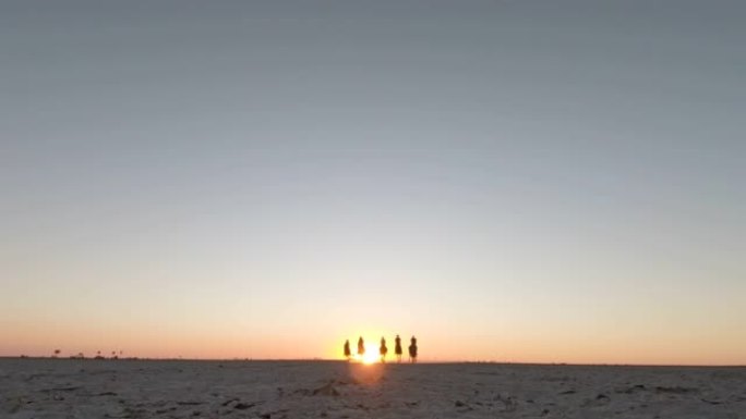 一群骑马者的慢动作低角度视图，在日落时分在Makgadikgadi盐锅的广阔空间中奔跑
