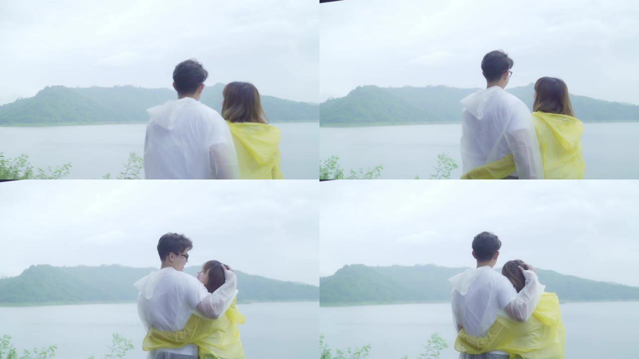 慢动作 -- 年轻的亚洲甜蜜夫妇穿着雨衣站在湖边，享受浪漫的雨水时光，感到快乐。生活方式夫妇在雨天享