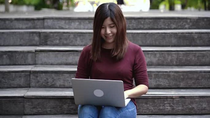 4k镜头亚洲迷人女性休闲服装坐在木楼梯上，在户外公园与科技笔记本电脑一起工作，生活方式和休闲概念