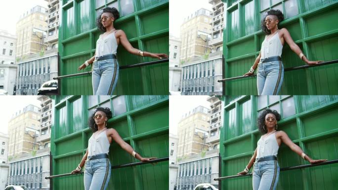 时尚的年轻黑人女性警告圆形太阳镜、吊带背心和牛仔裤靠在城市街道的扶手上，低角度