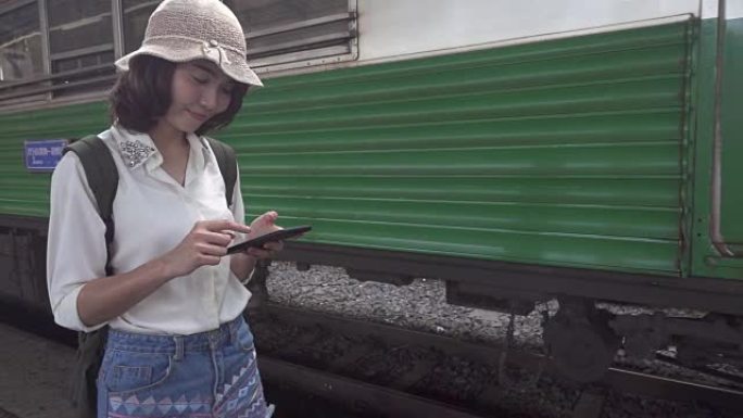 慢动作-旅行者背包客亚洲女人在泰国曼谷旅行。快乐的年轻女性使用智能手机在火车站聊天、阅读和发短信。