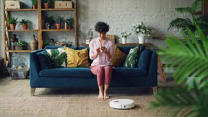 开朗的年轻女士正在打开机器人胡佛，坐在沙发上享受自动清洁，用智能手机放松。现代科技与青年观念。