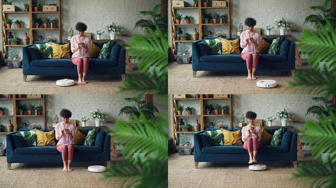 开朗的年轻女士正在打开机器人胡佛，坐在沙发上享受自动清洁，用智能手机放松。现代科技与青年观念。
