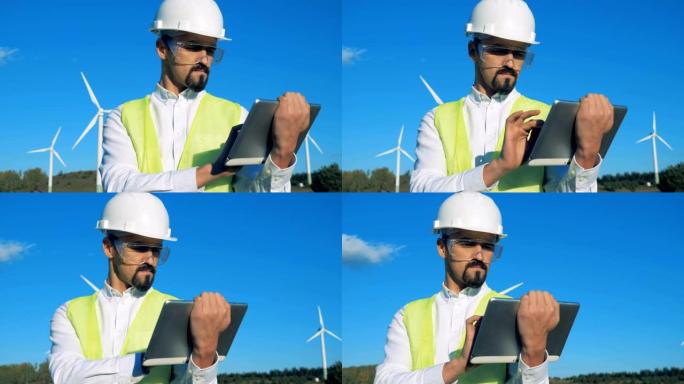 工人站在白色风力涡轮机附近，在平板电脑上打字。风车，绿色能源概念。