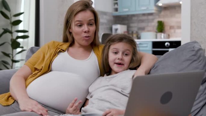 30多岁可爱的甜美母亲和学龄前儿童惊喜地看着笔记本电脑。漂亮的孕妇和可爱的小女儿坐在沙发上聊天，聊天