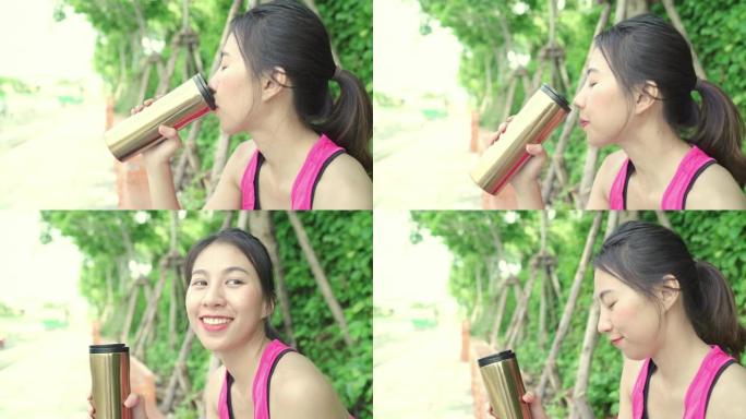 健康美丽的年轻亚洲跑步者妇女喝水，因为在城市公园的街道上跑步后感到疲倦。生活方式适合和活跃的女性在城