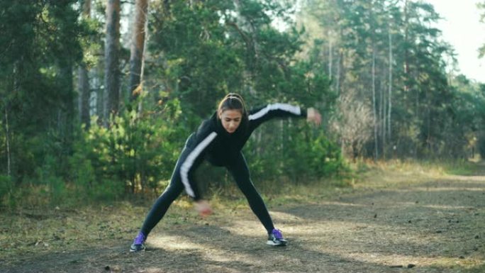 迷人的年轻女运动员在阳光明媚的秋天穿着现代黑色运动服在公园做运动。苗条的女孩正弯腰向前移动手臂。