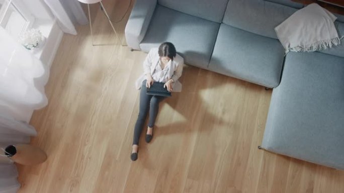 年轻女子坐在地板上，在笔记本电脑上工作或学习。舒适的客厅配有现代室内、灰色沙发和木地板。顶视图摄像机