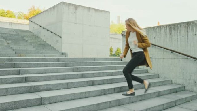 开朗快乐的年轻女子在走上楼梯时积极跳舞。她穿着一件棕色长外套。在商务中心旁边的城市混凝土公园拍摄的场