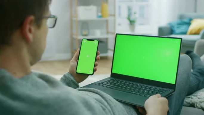 家里的年轻人在带有绿色模拟屏幕的笔记本电脑上工作，同时拿着带有色度键显示的智能手机。他坐在舒适客厅的