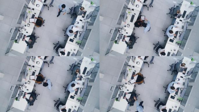 空中自上而下的无人驾驶飞机拍摄了繁忙的公司办公室，在台式计算机上工作的商人和女商人身上排了两排。明亮