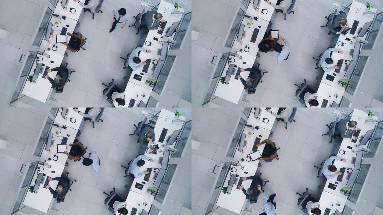空中自上而下的无人驾驶飞机拍摄了繁忙的公司办公室，在台式计算机上工作的商人和女商人身上排了两排。明亮