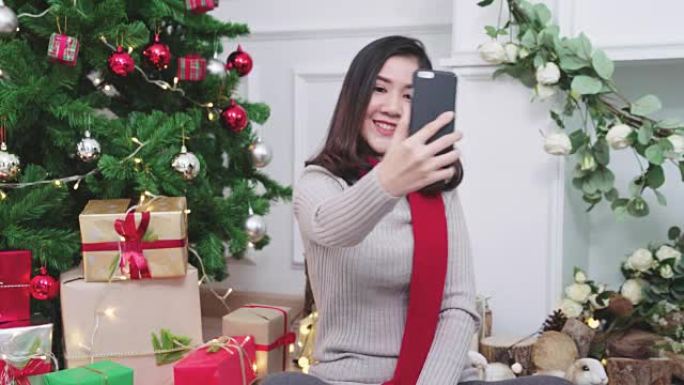 年轻迷人的亚洲女性使用智能手机与她的朋友在客厅进行视频聊天，对圣诞树背景感到满意。