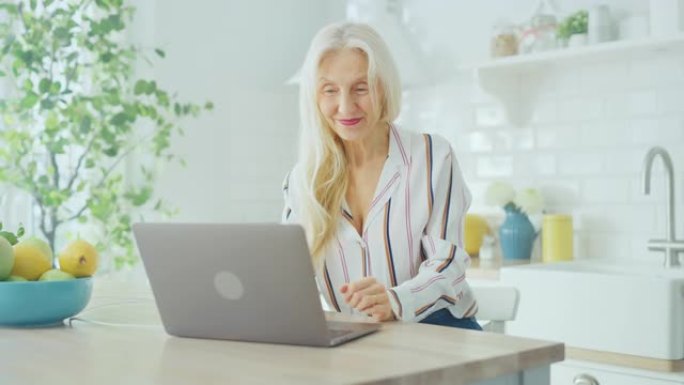 美丽的高级女人使用笔记本电脑，在阳光明媚的厨房里喝一杯咖啡或茶。祖母通过互联网和社交媒体与孩子建立联
